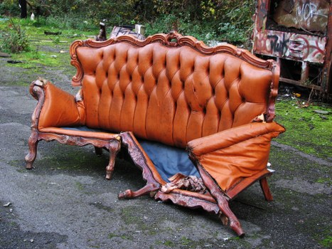 A worn-out, vintage-style sofa with ornate wooden carvings along the frame and a tufted, leather-upholstered backrest in a warm brown tone. Part of the sofa is damaged, with the fabric torn and the cushioning exposed, revealing a roughly cut blue material underneath. The armrest on the right side is dented and misshapen, indicating significant wear. The sofa is positioned outdoors on a paved driveway or path surrounded by greenery, with trees and grass visible in the background. To the right, there is a rusty, graffiti-covered metal structure or container, and the overall environment appears to be a yard or alleyway where rubbish removal services might collect bulky waste. The lighting is natural, softly illuminating the scene, emphasizing the texture of the damaged upholstery and the weathered wood. Commercial Waste Beaconsfield may be contacted for disposing of such bulky furniture waste in the Beaconsfield area, simplifying rubbish removal for local households or businesses.