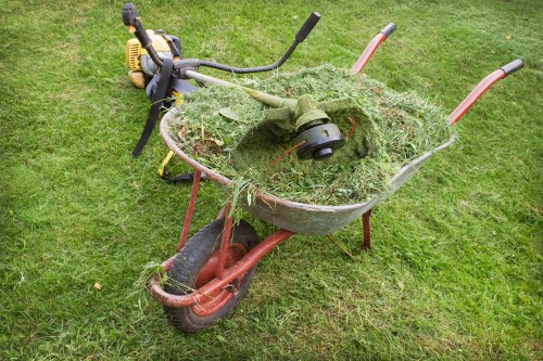 A red plastic wheelbarrow with a black rubber tire, filled with grass clippings, weeds, and garden debris, is positioned on a well-maintained grassy lawn, with a yellow and black petrol-powered grass trimmer resting nearby. The grass appears freshly cut, with clippings accumulated in the wheelbarrow, indicating recent garden clearing activity. The background features more green grass, suggesting an outdoor setting typical for garden maintenance in Beaconsfield, as managed by Commercial Waste Beaconsfield. The scene depicts typical garden waste ready for disposal or collection, with the wheelbarrow in the foreground, slightly tilted to one side, and the grass trimmer leaning against it. The natural outdoor environment emphasizes the context for rubbish removal services related to garden waste management in the local area, with no other objects or structures visible in the image.
