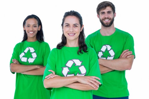 Three individuals wearing bright green T-shirts with white recycling symbols on the front, standing side by side outdoors against a plain background. The woman in the center has dark, shoulder-length hair and is smiling directly at the camera with her arms crossed. To her left, a woman with long dark hair also smiles and crosses her arms, while to her right, a bearded man with short dark hair and a friendly expression also has his arms crossed. The clothing and posture suggest they are part of a waste management or rubbish removal team, emphasizing professionalism and environmental awareness. The bright green shirts with recycling logos underline their association with eco-friendly rubbish collection services, suitable for a website about commercial waste removal in Beaconsfield. The overall scene conveys a clean, organized, and approachable team ready to assist with waste disposal and rubbish clearance tasks, in a manner consistent with local service providers such as Commercial Waste Beaconsfield.