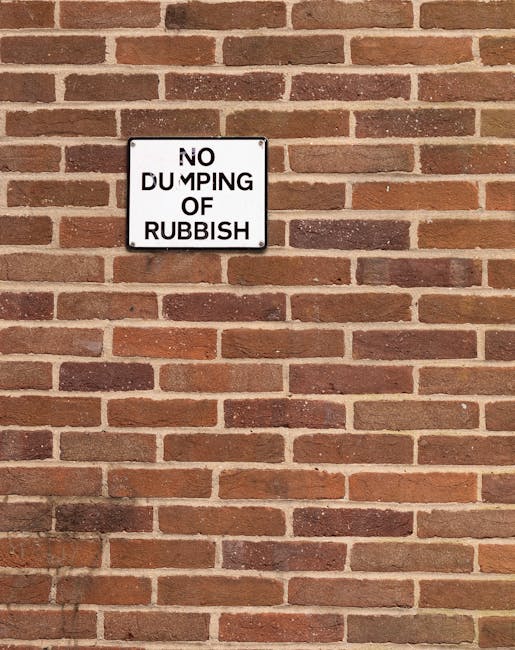 A close-up of a rectangular white metal sign mounted on a red brick wall. The sign displays the message 'NO DUMPING OF RUBBISH' in bold black uppercase letters, with the word 'DUMPING' partially obscu